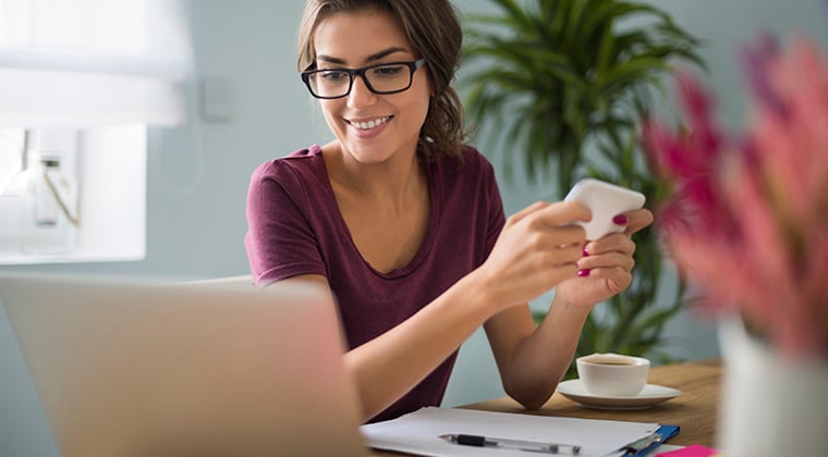 Woman sitting at desk holding mobile phone and looking at laptop computer.