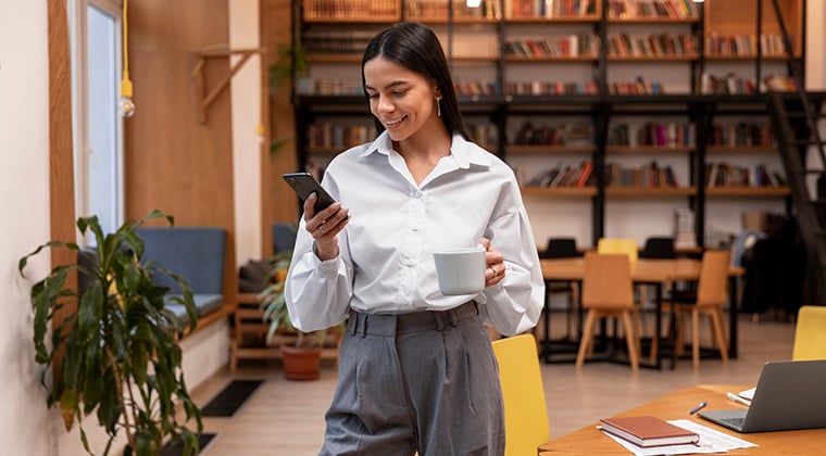 A woman in her office looks at a phone and smiles. 